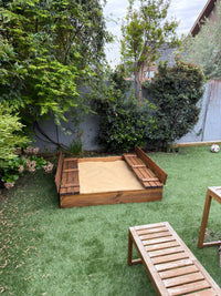 Arenero de madera para niños de 150 × 150 cm instalado en el pasto del jardín, con arena y las tapas abiertas como bancas